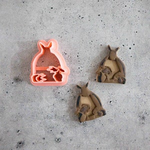 May include: Two cookie cutters, one pink and one brown, shaped like a Totoro character from the animated film My Neighbor Totoro. The cutters are on a gray textured surface.