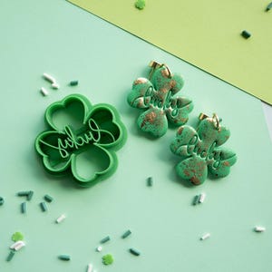 May include: A green clover-shaped cookie cutter and a pair of matching clover-shaped earrings. Both the cutter and the earrings feature the word "lucky" embossed in script. The earrings have a textured green finish with metallic gold accents.