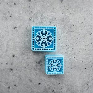 Pode incluir: Dois cortadores quadrados de cor azul-claro com um padrão de filigrana simétrico e intrincado dentro de uma moldura. Os objetos são construídos com um material sólido e apresentam um design detalhado em relevo para criar impressões.