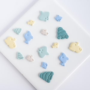 May include: An assortment of pastel-colored sea creature-shaped decorations, including seahorses, fish, and stingrays. The decorations are in shades of blue, yellow, and pink, arranged on a white surface. These could be used for crafting or decorating.