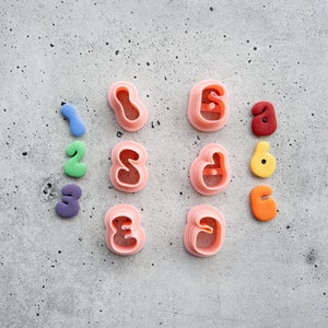 May include: A set of pink plastic cookie cutters shaped like the numbers 1, 2, 3, 4, 5, and 6. The cutters are arranged in two rows of three. There are also six small, colorful fondant shapes in the image, representing the numbers 1 through 6.