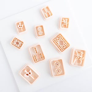 Puede incluir: Una colecci&oacute;n de cortadores de galletas en forma de fichas de Mahjong de color marr&oacute;n claro. Cada cortador presenta un dise&ntilde;o diferente, incluyendo bamb&uacute;, c&iacute;rculos y caracteres chinos. Los cortadores est&aacute;n dispuestos sobre una superficie blanca.