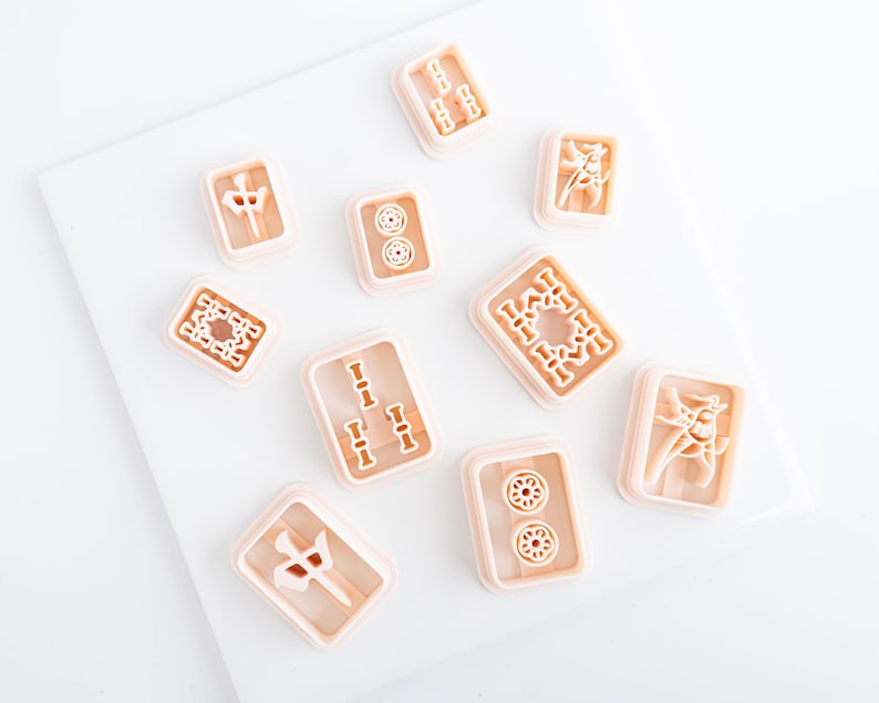 Puede incluir: Una colecci&oacute;n de cortadores de galletas en forma de fichas de Mahjong de color marr&oacute;n claro. Cada cortador presenta un dise&ntilde;o diferente, incluyendo bamb&uacute;, c&iacute;rculos y caracteres chinos. Los cortadores est&aacute;n dispuestos sobre una superficie blanca.