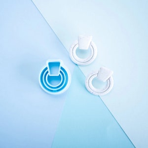 Puede incluir: Dos pares de pendientes sobre un fondo azul y azul claro. Un par es azul y tiene forma de anillo con tres capas. El otro par es blanco con un acabado moteado y tiene forma de anillo con un óvalo pequeño en el centro.