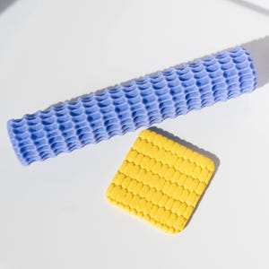 May include: A blue textured roller and a yellow textured square are displayed on a white surface. The roller is cylindrical and has a repeating pattern of rounded shapes. The square has a grid-like pattern.