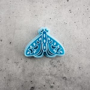 May include: A light blue plastic cookie cutter shaped like a moth with intricate cut-out details on the wings, including crescent moon and circular patterns. The object features a raised border and a recessed design for creating impressions in dough.