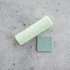 May include: A light mint green cylindrical roller tool and a small square tile, both featuring a raised, intricate leaf-like texture pattern. The roller is elongated, while the tile is compact with rounded corners, showcasing a matching design.