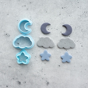 Puede incluir: Un conjunto de cortadores de galletas y formas de fondant azules y grises. Los cortadores incluyen una luna creciente, una nube y una estrella. Las formas de fondant son una luna creciente, una nube y una estrella.