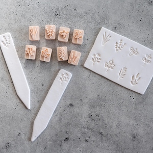 May include: A set of eight small peach-colored square stamps featuring various botanical designs, two white pointed plant markers with embossed floral patterns, and one white rectangular slab with eight matching botanical impressions.