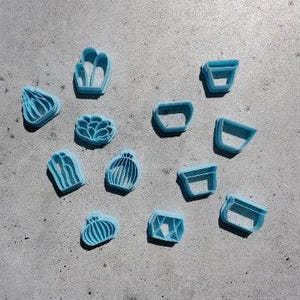 May include: A collection of twelve light blue plastic cookie cutters in various shapes, including several cactus and succulent designs alongside multiple geometric pot shapes, arranged in a scattered pattern on a flat surface.