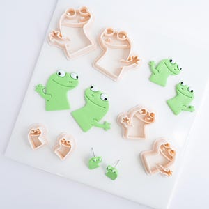 May include: A selection of frog-shaped cookie cutters and fondant decorations. The cutters are a light peach colour, and the frogs are bright green. The frog shapes include various sizes and designs, ideal for baking and crafting projects.
