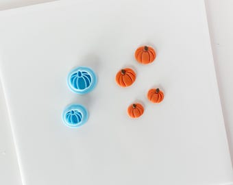 May include: Two light blue plastic pumpkin-shaped cookie cutters are positioned next to four small, orange, molded pumpkin decorations with brown stems. The items are arranged on a plain white surface, showing the tools and the finished shapes.