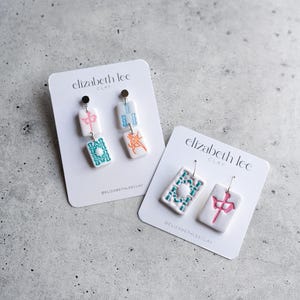 Puede incluir: Dos pares de pendientes de arcilla blanca con dise&ntilde;os coloridos. Un par tiene detalles en rosa, azul y naranja, mientras que el otro presenta dise&ntilde;os en verde azulado y rosa. Cada pendiente est&aacute; en una tarjeta blanca con el texto "elizabeth lee CLAY".