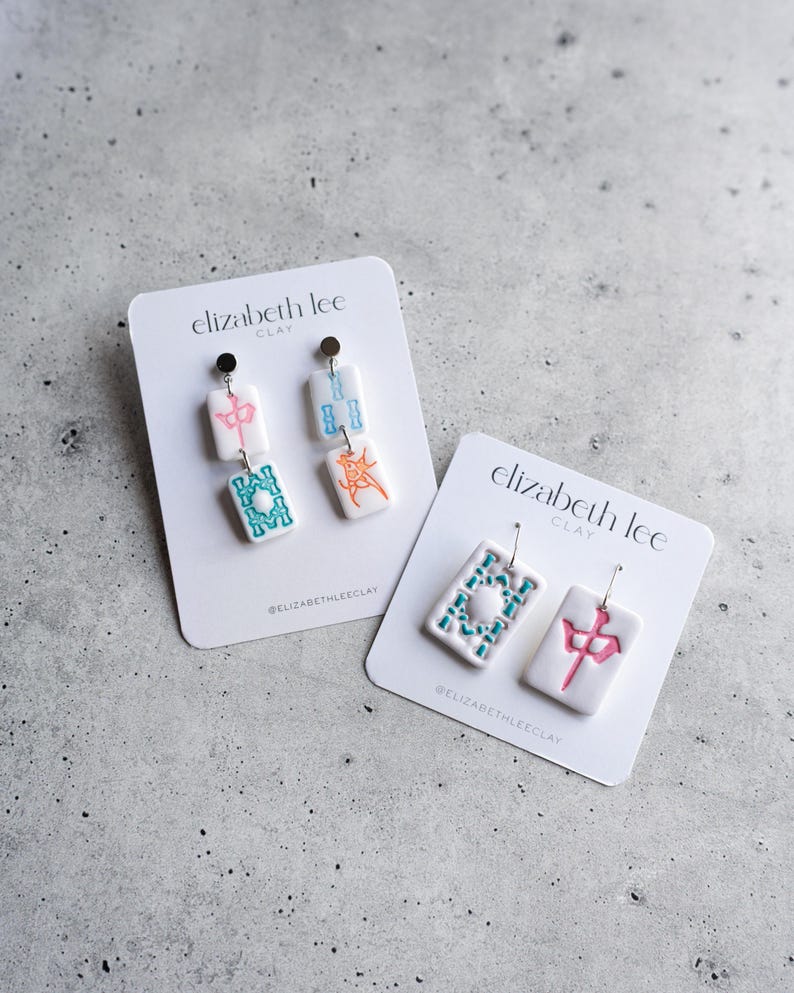 Puede incluir: Dos pares de pendientes de arcilla blanca con dise&ntilde;os coloridos. Un par tiene detalles en rosa, azul y naranja, mientras que el otro presenta dise&ntilde;os en verde azulado y rosa. Cada pendiente est&aacute; en una tarjeta blanca con el texto "elizabeth lee CLAY".