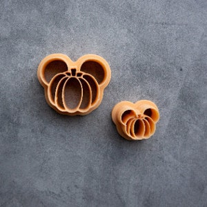 May include: Two orange plastic cookie cutters shaped like pumpkins. The larger cutter has two rounded ears on top and the smaller cutter is a simple pumpkin shape.