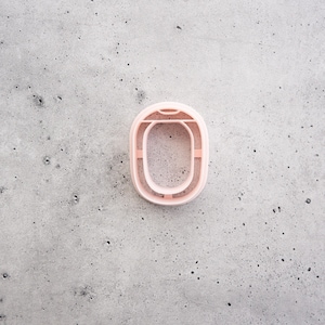 May include: A light pink, oval-shaped cookie cutter with a smooth, rounded frame and a central open space. The cutter features a thin, raised edge designed for cutting dough into a specific shape, showcasing a clean and simple construction.