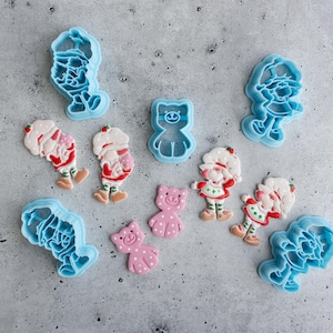 May include: A set of nine blue plastic cookie cutters featuring various characters, including a bear, a pig, and a girl with a hat. The cutters are arranged on a gray textured surface.
