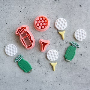 Puede incluir: Un juego de cortadores de galletas de plástico rosa y verde con forma de bolas de golf, palos de golf y una bolsa de golf. Los cortadores están sobre una superficie gris texturizada.