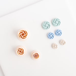 以下が含まれることがあります： さまざまな色の小さな装飾ボタンの詰め合わせ。ボタンは幾何学的な結び目のようなデザインです。色は、ライトオレンジ、ライトブルー、ライトグリーン、ベージュなどです。ボタンは白い表面に配置されています。