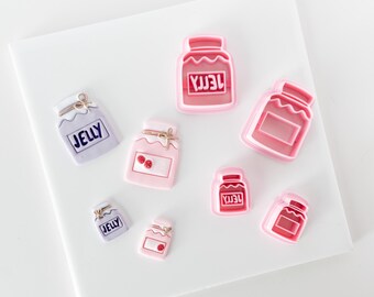 May include: A collection of jelly jar shaped cookie cutters and corresponding clay or fondant decorations. The pink plastic cutters feature a jar outline with the word JELLY embossed. The decorations include light purple and pink jars with JELLY text.