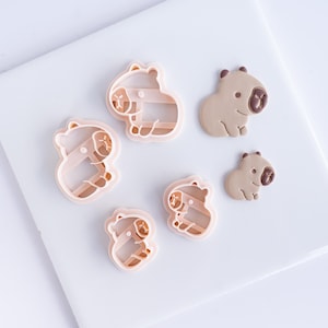 Puede incluir: Un conjunto de cortadores de galletas y galletas con forma de carpincho. Los cortadores son de color rosa claro, y las galletas son de color marrón claro con detalles marrones. Los cortadores son de plástico, y las galletas son de masa.