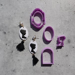 May include: Two pairs of earrings with black tassels and white oval-shaped charms featuring a black silhouette of a woman's head.  Five purple plastic cookie cutters in various shapes, including a circle, a rectangle, and a woman's head.