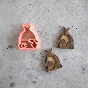 May include: A peach-colored plastic cookie cutter shaped like a kangaroo with a joey in its pouch, accompanied by two finished brown clay kangaroo figures that mirror the shape and design of the cutter, showing the same animal silhouette.
