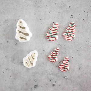 May include: Two white plastic Christmas tree-shaped cookie cutters and four small, flat Christmas tree-shaped decorations. The decorations are white with red horizontal stripes and small green sprinkles scattered across the surface.