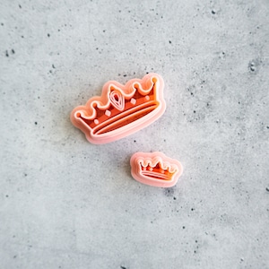 May include: Two pink plastic crown-shaped cookie cutters. The larger cutter is in the upper left corner of the image and the smaller cutter is in the lower right corner of the image.