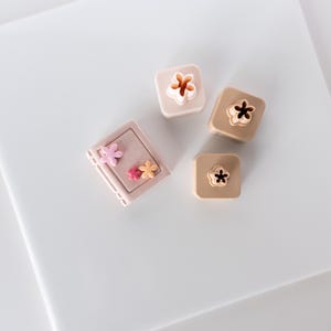 May include: Four small pink and brown boxes with flower designs. The boxes are arranged in a square shape on a white surface.