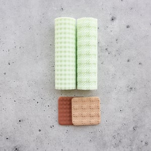 Pode incluir: Dois panos verde-claros enrolados com padrões texturizados e dois panos quadrados em tons de castanho. Os panos estão dispostos sobre uma superfície cinza salpicada. Os panos enrolados têm um padrão xadrez e diagonal.