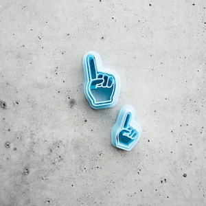 May include: Two blue plastic cookie cutters shaped like a raised fist with the index finger pointing up.