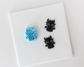 Könnte beinhalten: Ein hellblauer Ausstechform aus Kunststoff in Form eines Drachen mit spitzen Ohren und kleinen Flügeln, begleitet von zwei schwarzen Drachenanhängern mit grünen Augen und detaillierten Gesichtszügen, angeordnet auf einer weißen Fläche.