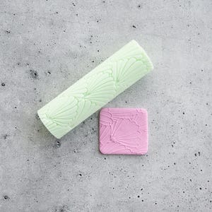 Pode incluir: Um rolo cilíndrico verde-claro com um padrão de conchas em relevo e um pequeno azulejo quadrado cor-de-rosa com o mesmo design de conchas gravado. Ambos os itens exibem um motivo repetitivo de conchas em forma de leque nas superfícies.