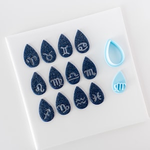 Op de afbeelding: Een verzameling van twaalf donkerblauwe, glinsterende druppelvormige stukjes, elk voorzien van een zilverkleurig sterrenbeeld. Daarnaast liggen twee lichtblauwe, druppelvormige plastic uitsteekvormpjes, waarvan er één een symbool bevat.