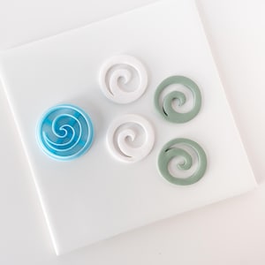 May include: A collection of decorative swirl-shaped objects in various colors. One is blue and white, two are white, and two are a muted green. The objects are arranged on a white surface, creating a clean and modern aesthetic.