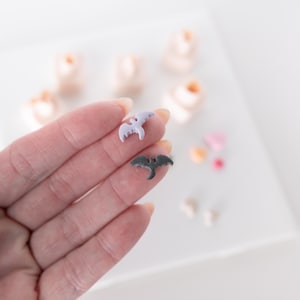 Op de afbeelding: Twee kleine vleermuisvormige bedels, één in licht lavendel en één in donker antracietgrijs, gehouden tegen een wazige achtergrond van extra kleine, lichtgekleurde en pastelkleurige decoratieve vormen op een witte ondergrond.