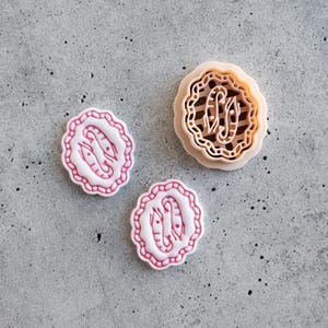 May include: Three cookie cutters in a top-down view. Two are white with pink shrimp designs and scalloped edges. The third is tan with a shrimp design and a striped interior. All are on a gray surface.
