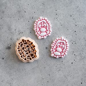 May include: Three crab-shaped cookie cutters and cookies. Two white cookies with red crab designs and scalloped edges. A beige cookie cutter with a crab design and a scalloped edge.
