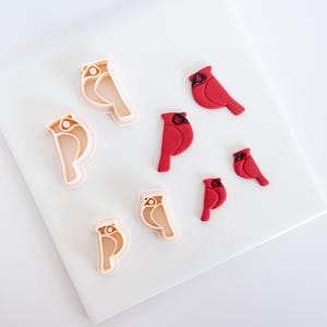 May include: A collection of cardinal bird-shaped cookie cutters and finished cookies. The cutters are light tan, while the cookies are bright red. The shapes are arranged on a white surface, showcasing the contrast between the tools and the finished product.