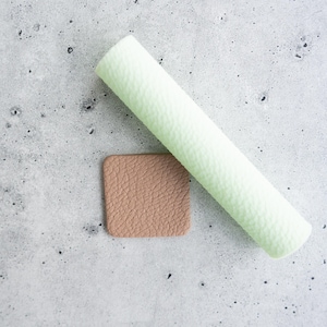 May include: A light green cylindrical roll of textured material positioned diagonally next to a small, square-shaped piece of tan material with a matching pebbled texture. Both items feature a consistent, grainy surface pattern throughout.