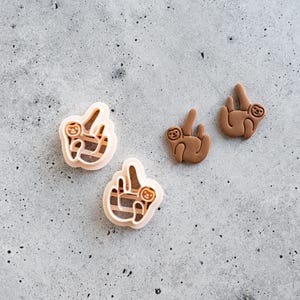May include: Two light peach-colored plastic sloth-shaped cookie cutters and two brown sloth-shaped cutouts. The cookie cutters feature an open frame design with internal details for the sloth's face and limbs, while the brown cutouts are solid.