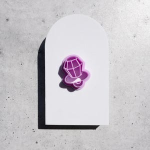 May include: A light purple plastic cookie cutter shaped like a baby pacifier with a faceted diamond-like top and a circular handle base. The cutter features a raised rim and a recessed center, designed for cutting dough into a specific shape.