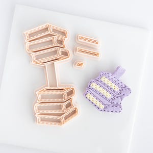 May include: A collection of book-shaped cookie cutters and a felt book-shaped item. The cookie cutters are in various sizes and shapes, with a light brown color and stitched details. The felt item is purple with yellow and white details.