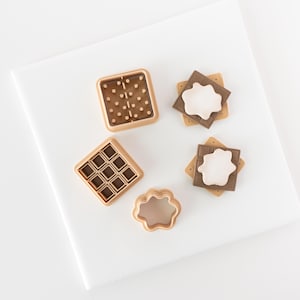 以下が含まれることがあります： 正方形、長方形、円形、穴の開いた異なるサイズの正方形2つの形をした、茶色のプラスチック製のクッキー型5個セット。型は白い表面の上に置かれています。