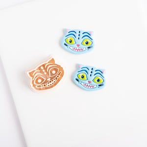 May include: Three cat-shaped cookie cutters. Two are light blue with yellow eyes, pink noses, and white teeth. One is a light orange outline with a detailed cat face design featuring stripes, large circular eyes, and a wide grin with visible teeth.