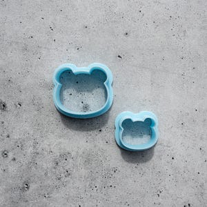 May include: Two light blue cookie cutters shaped like bear heads. The set includes one larger bear head cookie cutter and one smaller bear head cookie cutter, both featuring rounded ears and a simple outline design for baking shapes.
