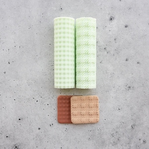 Pode incluir: Dois rolos cilíndricos com um padrão xadrez verde-claro e dois azulejos quadrados, um em castanho-avermelhado escuro e outro em bege claro, ambos com um design de grelha texturizado nas superfícies, dispostos em forma de ponto de exclamação.