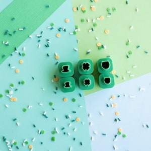 May include: A set of six green plastic cookie cutters with different St. Patrick's Day themed shapes, including a shamrock, a hat, and a pot of gold. The cutters are on a green and blue background with sprinkles.
