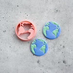 Op de afbeelding: Een roze plastic koekjesvorm in de vorm van de aarde naast twee koekjes in de vorm van de aarde. De koekjes hebben blauwe oceanen en groene continenten. De vorm is rond met de continenten uitgesneden.